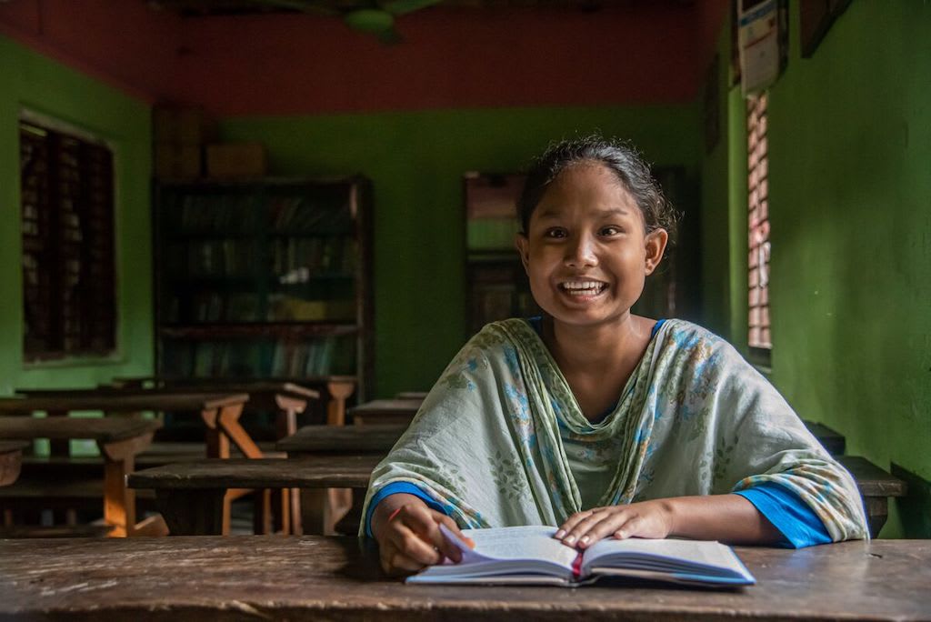 A girl wearing a loose green top smiles as she reads a book on a brown table in front of her.